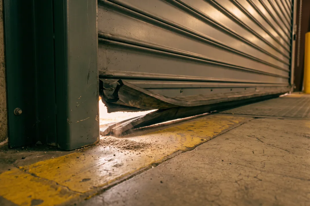 Primer plano a ras del piso de la esquina inferior de una puerta de andén enrollable de acero gris en un almacén: el burlete de goma está roto y la luz del día se cuela por el hueco bajo la puerta.