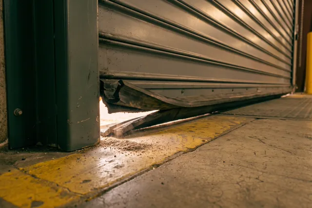 The bottom corner of a warehouse dock door, its torn rubber seal leaving a gap where daylight shows under the door.